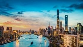 Panoramic cityscape of Shanghai at dusk, showing the Huangpu River curving through the city with illuminated skyscrapers in the Pudong financial district, including the Shanghai Tower and surrounding high-rise buildings. The scene highlights modern urban development, infrastructure, and economic growth in China, reflecting a key Asia-Pacific market often associated with international trade, regulatory compliance, and market access for global businesses.