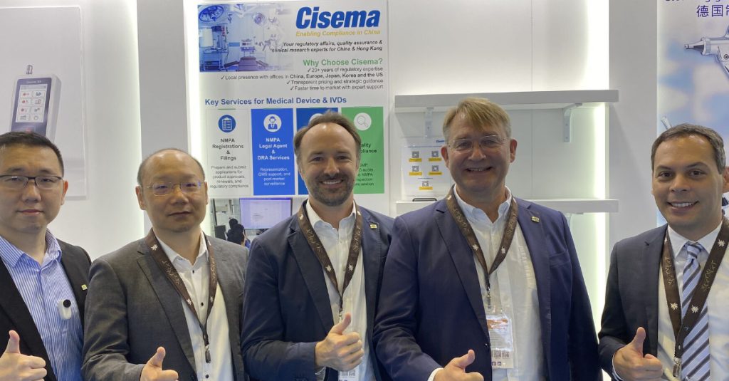 Five professionals stand side by side at a trade show booth branded “Cisema – Enabling Compliance in China.” They are wearing business attire and conference badges, smiling and giving thumbs-up gestures toward the camera. Behind them, a vertical display highlights regulatory consultancy services for medical devices and IVDs, including NMPA registrations, legal agent services, quality compliance, and market access support. The booth design features clean white panels, blue and green icons, and messaging focused on regulatory compliance, product registration, and clinical and quality expertise in China and the Asia-Pacific region. The scene conveys collaboration, expertise, and professional support for companies entering or expanding in regulated China and Asia-Pacific markets.